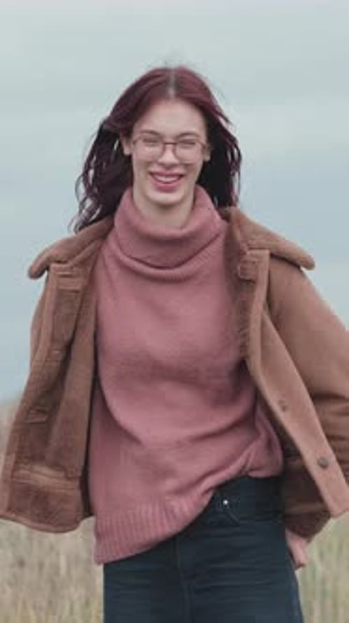 Smiling teenage girl wearing pink turtleneck sweater and brown coat standing outdoors.