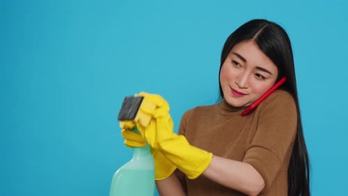 Asian Housekeeper Using Chemical Detergent and Duster to Clean House While Talking at Mobile Phone