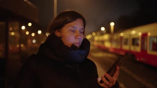 Frau in schwarzer Jacke benutzt nachts ein Telefon auf dem Bahnsteig einer S-Bahn