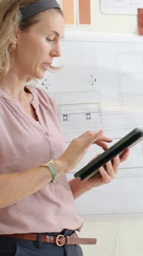Vertical video: Camera panning woman in pink top checking plans, swatches, tapping tablet at studio