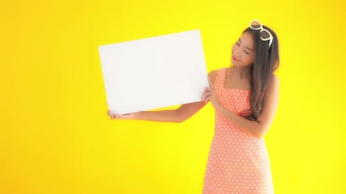 Young Asian woman in summer dress with sunglasses on head holds up plain white sign board and smiles