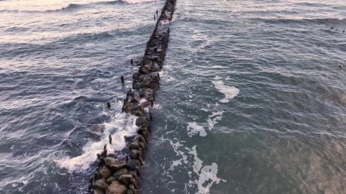 Quebra-mar que se estende até o mar cercado por ondas e costa rochosa ao pôr do sol