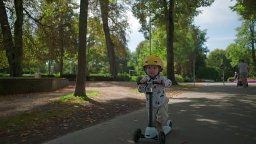 Little child with yellow helmet riding scooter along tree-lined path in park, moving forward with