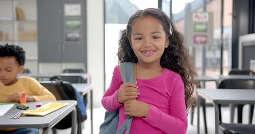 In school classroom, young biracial girl holding a backpack smiles