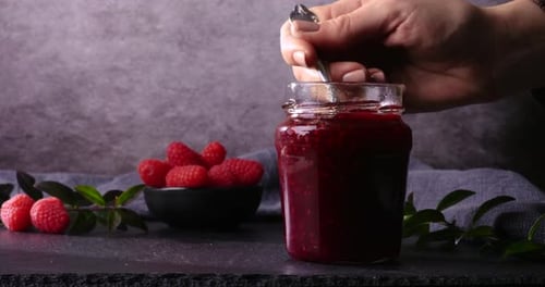 A close-up shot of a spoon lifting a rich scoop of raspberry jam from a jar. Fresh raspberries and g