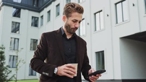 Man in a Brown Blazer Checks His Phone While Holding a Coffee Outside Office