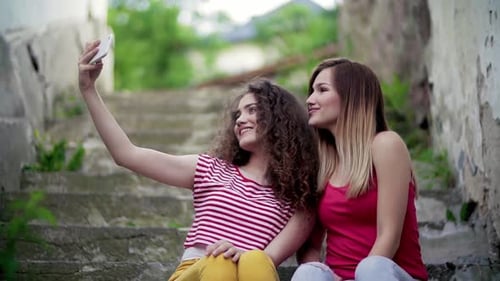 Teenage Female Friends Sitting Outdoors In Town, Taking Selfie With Smartphone.