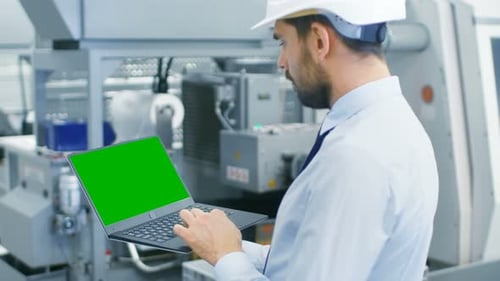 Industrial Engineer in Hard Hat Works on Green Mock-up Screen Laptop While Standing in the Middle o