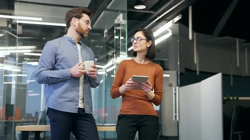 Two colleagues engaged in friendly conversation in modern office setting. Man holding coffee, woman