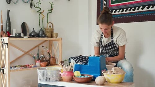 A female potter in jeans and an apron works on a potter's wheel, making a ceramic pot from clay