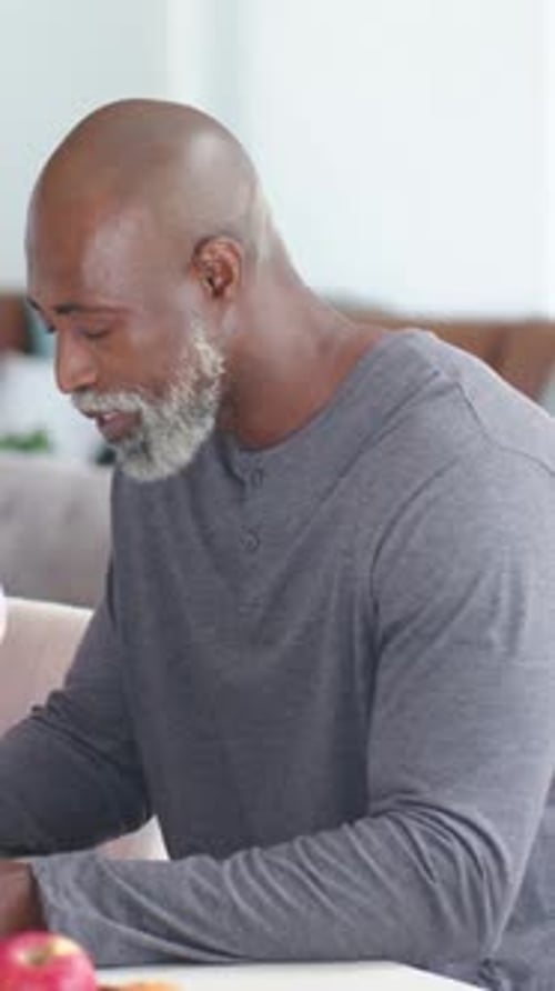 Vertical video: Noticing fruit, senior in grey henley leaning, moving fruit on white table at home