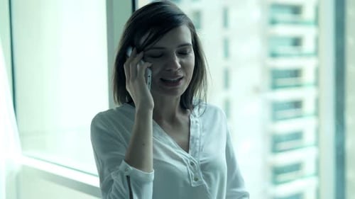 Young businesswoman chatting on cellphone by office window overlooking cityscape