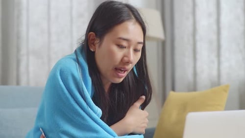 Close Up Of Sick Asian Woman With Blanket Holding Medicine To The Video Call With Doctor On A Laptop