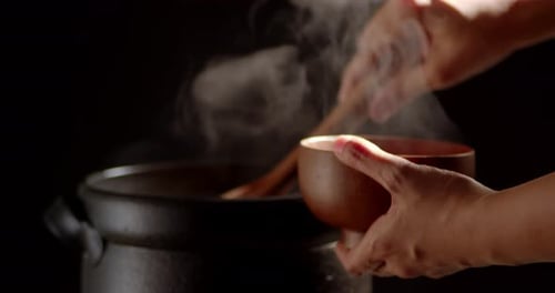 the cook is cooking food in a boiling clay pot with steam rising and using a wooden ladle