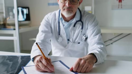 Portrait of Male Doctor in Lab Coat Sitting at Desk, Filling Medical Documents