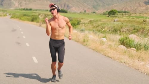 Shirtless Man in Sunglasses and Headphones Runs Smoothly Along a Scenic Road