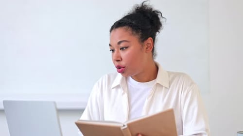 Woman reads book while using laptop indoors