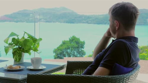 A Young Man on Vacation Sits in Hotel Cafe Terrace with View of Sea
