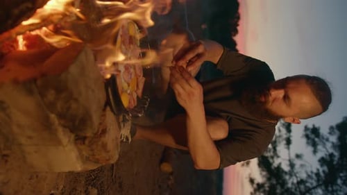 A Young Bearded Man Cooks Food on a Bonfire in a Frying Pan on the River Bank at Sunset