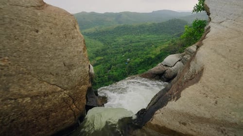 Rauschendes Wasser fließt vom Fluss in den Wasserfall über Felsen, grüne Hügel umgeben das Gebiet. Klare Sicht auf