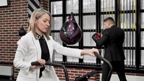 Couple of Businessmen Man and Woman Play Sports on an Exercise Bike and a Boxing Ring in Suits