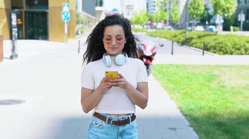 Happy Casual Beautiful Woman in Pink Glasses with Headphones Texting on Smartphone While Walking in