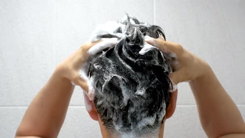 Male hand washes his head with shampoo and foam on gray background close-up, back view.