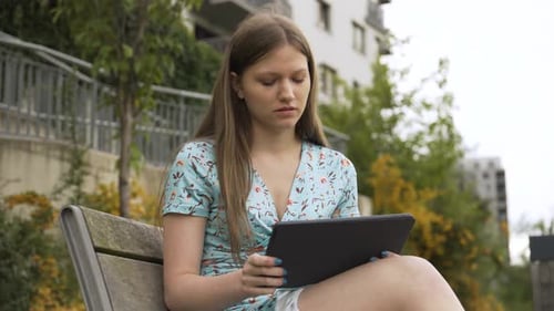 A Beautiful Young Caucasian Woman Works on a Tablet As She Sits on a Bench in a Suburban Area