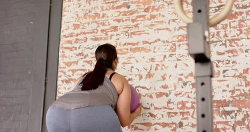 Woman retrieving medicine ball then repeating throwing and catching on brick wall for fitness