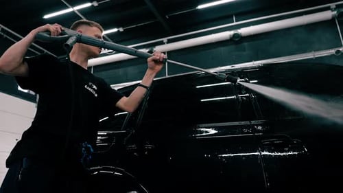 A male car wash employee applies car wash detergent to a black luxury car using spray gun in the car