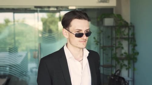 handsome, stylish young Caucasian man in business attire is putting on sunglasses in a trendy office