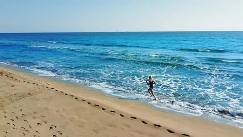 Drone view of woman in black swimsuit running on sand along sea coast.