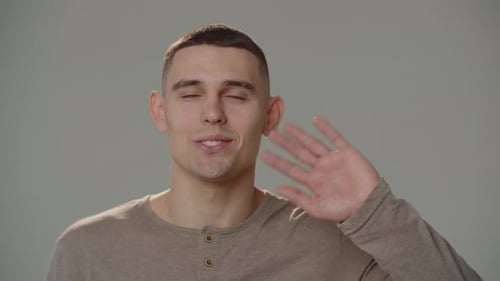 Smiling Man Greets and Waves His Hand to the Camera Frontal Shot of a Young Smiling Man in Front of