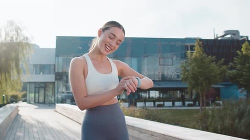 Athletic Runner Woman Looks at Smartwatch Device Fitness Bracelet Checking Heart Rate in City Park