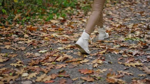 Female in tight beige leggings and white boots walks quickly by the path in the park.