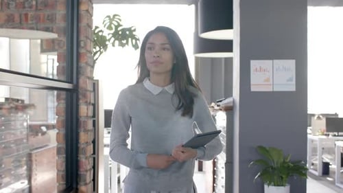 Biracial casual businesswoman walking through office with tablet, slow motion
