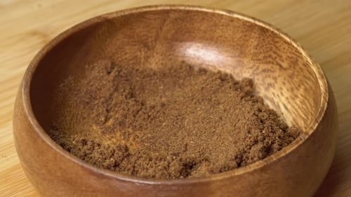 Spoon picks up dried nutmeg spice from wooden bowl and rotating on wooden board. Dry seasoning