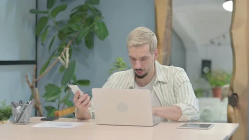 Young Man Uses Laptop and Smartphone at Desk