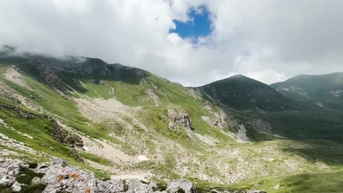 A mountain landscape featuring rocky slopes, grassy areas under a cloudy sky. The mountain peaks are