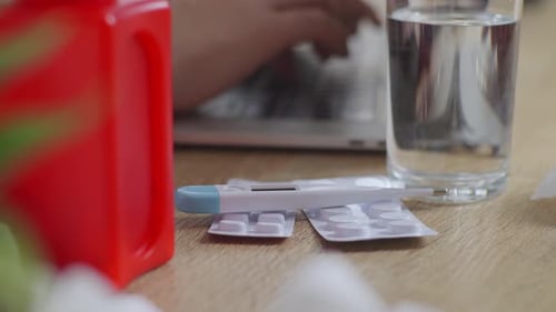 Varios medicamentos recetados y un termómetro sobre la mesa, mano de empresario escribiendo en la computadora portátil