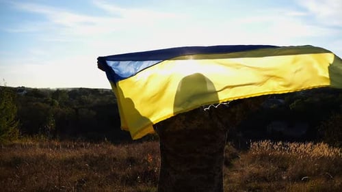 Military Man in Uniform Stands with Raised Over Head Ukraine Flag Against Sunlight at Background