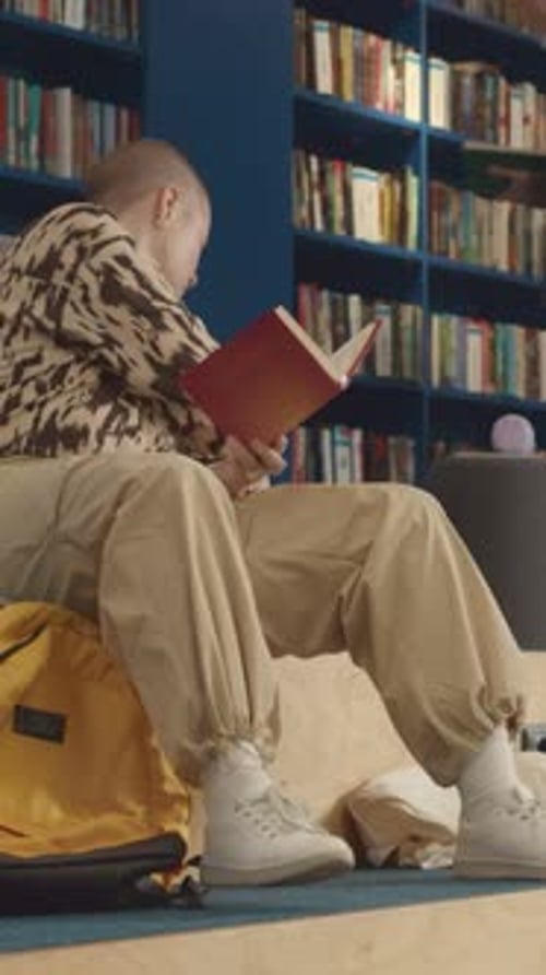 Female Student with Shaved Head Reading Textbook in College Library