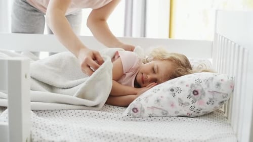 Young mother kisses her little daughter that lying on the bed and going to sleep indoors at home