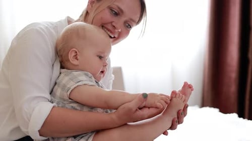 Muttertag. Lächelnde junge Mutter spielt mit den Beinen eines kleinen Mädchens auf dem Bett im Schlafzimmer zu Hause. Liebevolle Mama