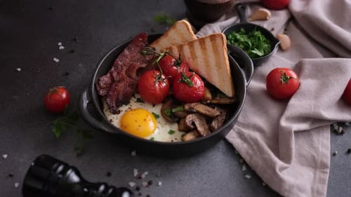 Une femme met un délicieux plat de petit déjeuner sur fond foncé, bacon frit, œufs, champignons et tomates