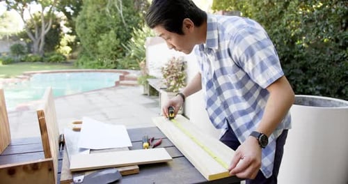 Measuring wooden plank, asian man working on DIY project in backyard workshop