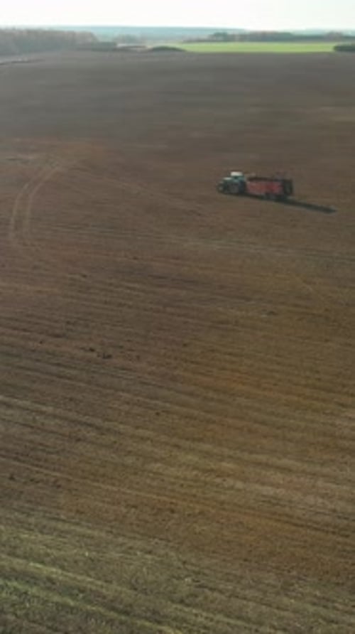 Vertical Wide Aerial View of a Tractor with a Trailer Driving Through a Vast Brown Field