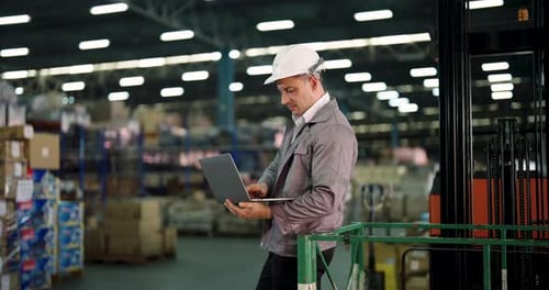 caucasian male warehouse staff working with laptop near stacked boxes checking stock status beside