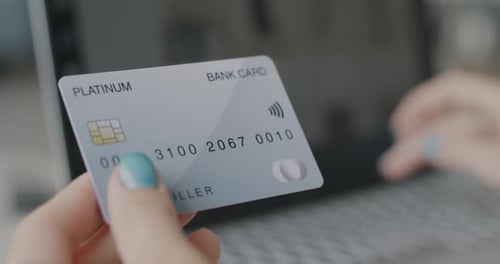 Nahaufnahme einer Frau, die eine Bankkarte in der Hand hält und drinnen am Schreibtisch mit Laptop tippt