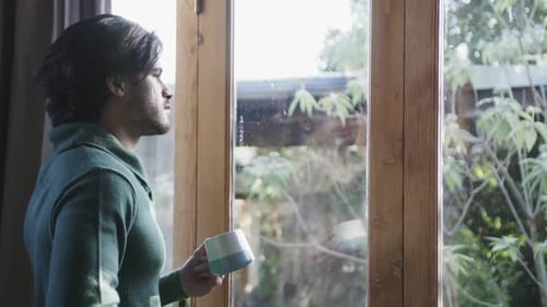 Thoughtful caucasian man with cup of coffee and looking out window at home, copy space, slow motion